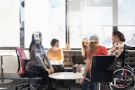 A diverse group of women having a discussion in a bright, modern office. One participant is using a wheelchair, and another has a laptop open.